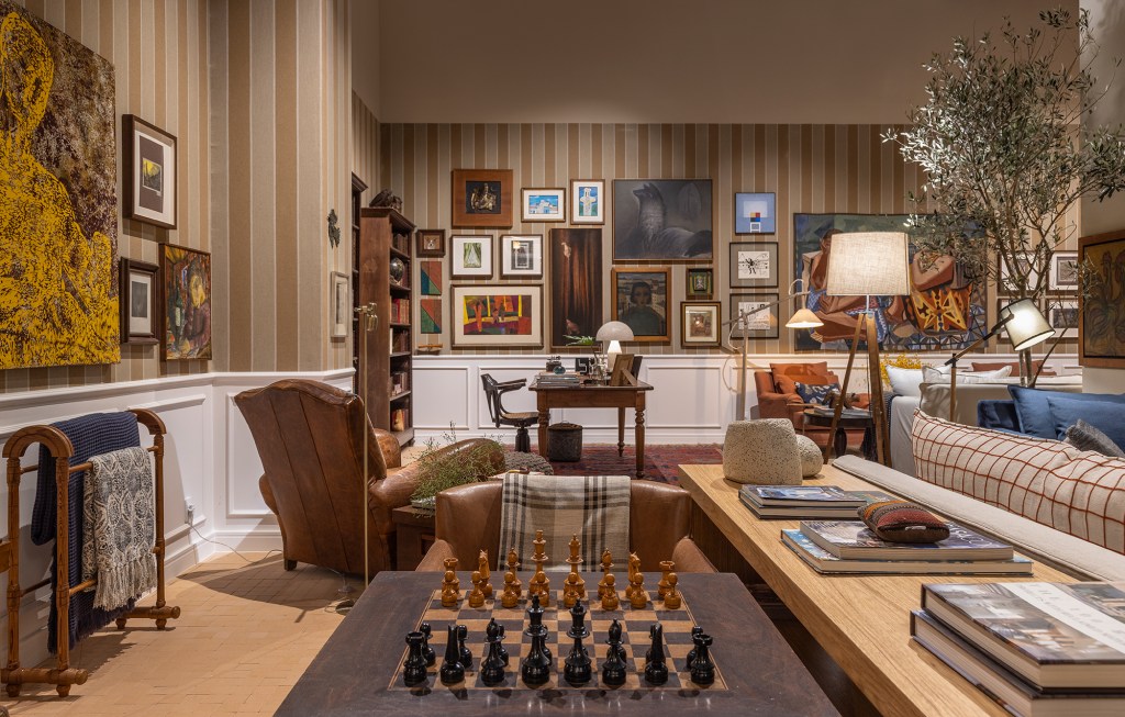 Com TV de 136 polegadas, sala de 125 m² é repleta de lembranças de família. Projeto de Felipe de Almeida para a CASACOR SP 2023. Na foto, sala com sofá, poltrona, quadros e jogo de xadrez.