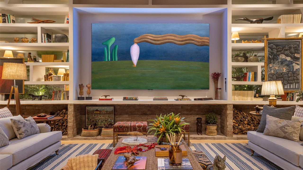 Com TV de 136 polegadas, sala de 125 m² é repleta de lembranças de família. Projeto de Felipe de Almeida para a CASACOR SP 2023. Na foto, sala com sofá, quadro, tapete, estante.