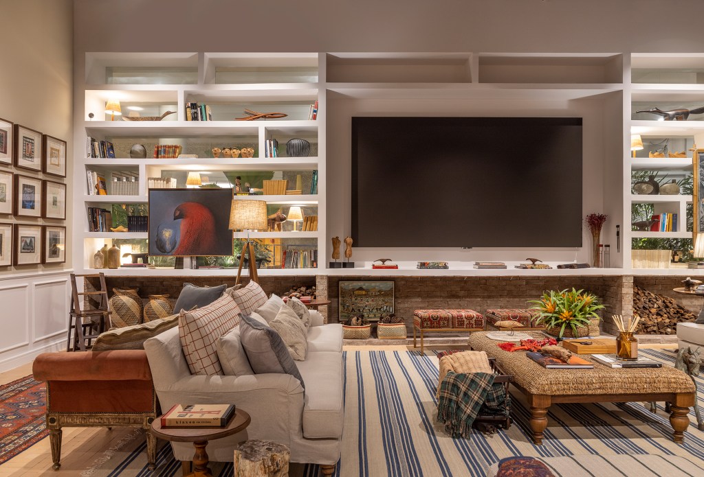 Com TV de 136 polegadas, sala de 125 m² é repleta de lembranças de família. Projeto de Felipe de Almeida para a CASACOR SP 2023. Na foto, sala com sofá, quadro, tapete, estante.