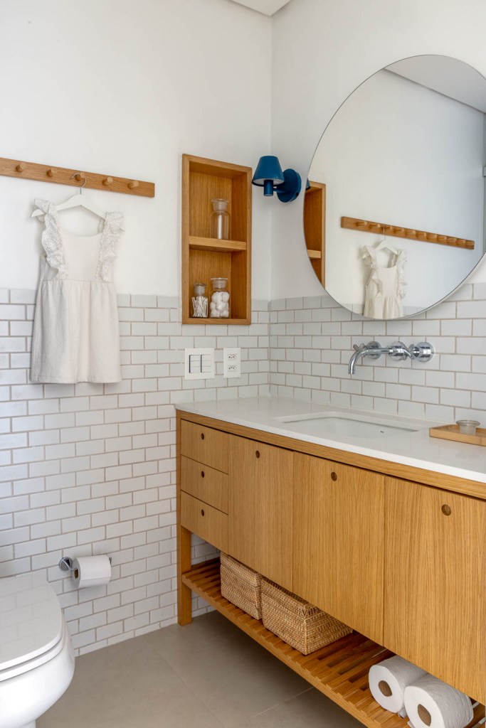 Banheiro com bancada branca, espelho redondo e parede com tijolinhos.