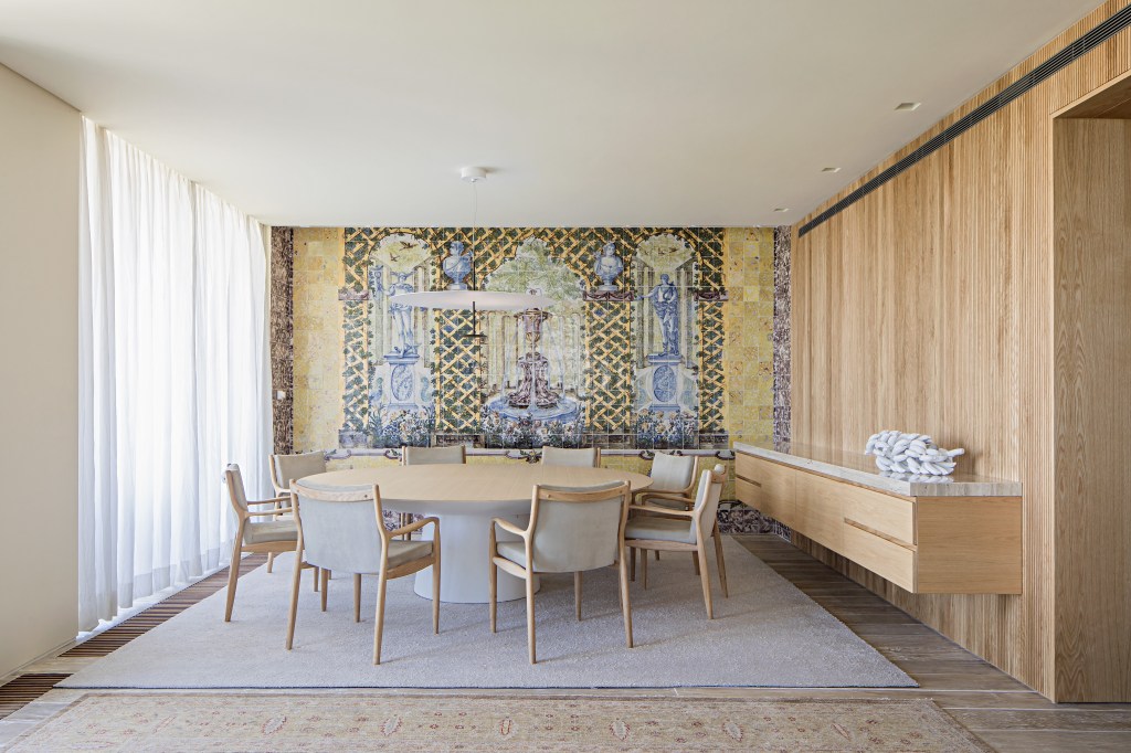 Sala de jantar com mesa de madeira redonda, painel de azulejos antigos ao fundo e tapetes.