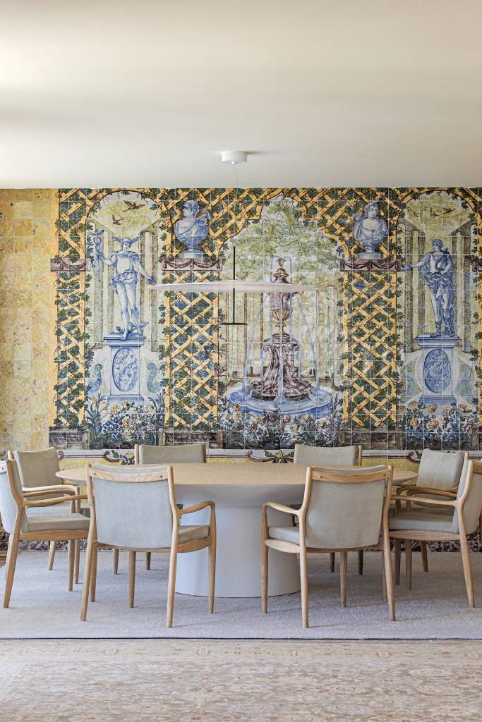 Sala de jantar com mesa de madeira redonda, painel de azulejos antigos ao fundo e tapetes.