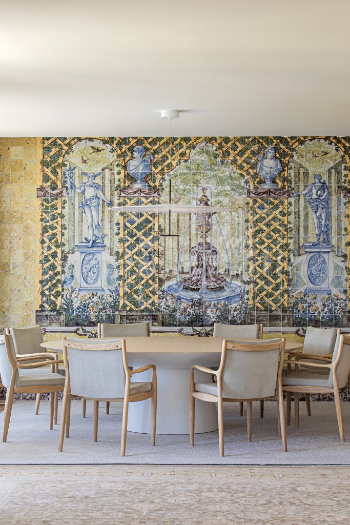 Sala de jantar com mesa de madeira redonda, painel de azulejos antigos ao fundo e tapetes.