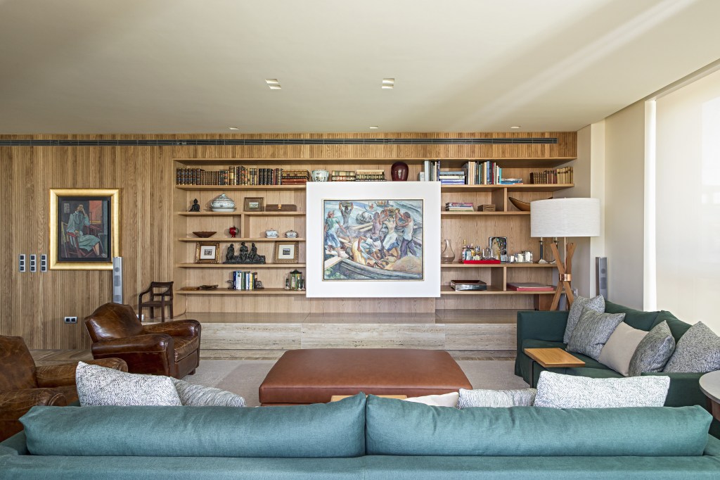 Sala de estar com estante de madeira com quadro que desliza, sofá verde, poltronas vintage de couro e tapetes.