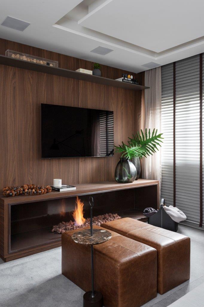 Sala de estar com parede revestida com painel de madeira, tv, lareira e pufe de couro.