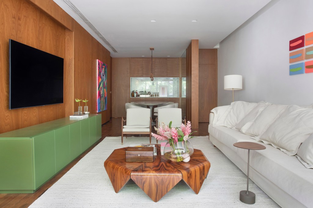Projeto de Bianca Da Hora. Na foto, sala de estar com rack verde, sofá branco, poltronas brancas, tapete off white e mesa de centro de madeira.
