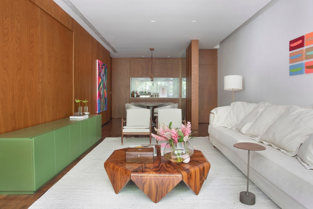 Projeto de Bianca Da Hora. Na foto, sala de estar com rack verde, sofá branco, poltronas brancas, tapete off white e mesa de centro de madeira.