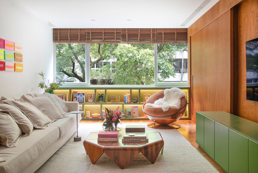 Projeto de Bianca Da Hora. Na foto, sala de estar com poltrona, tapete off white, mesa de centro de madeira e estante baixa com nichos verde.