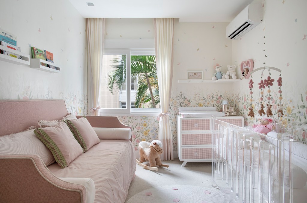 Projeto de Bianca Da Hora. Na foto, quarto de bebê com papel de parede floral, poltrona branca, sofá cama e berço de acrílico branco.