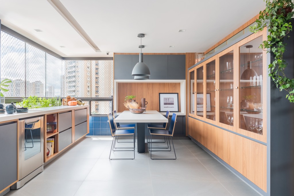 Projeto de Pietro Terlizzi. Na foto, varanda integrada com cozinha com cristaleira, mesa para refeições, piso de porcelanato e parede de azulejos azuis.