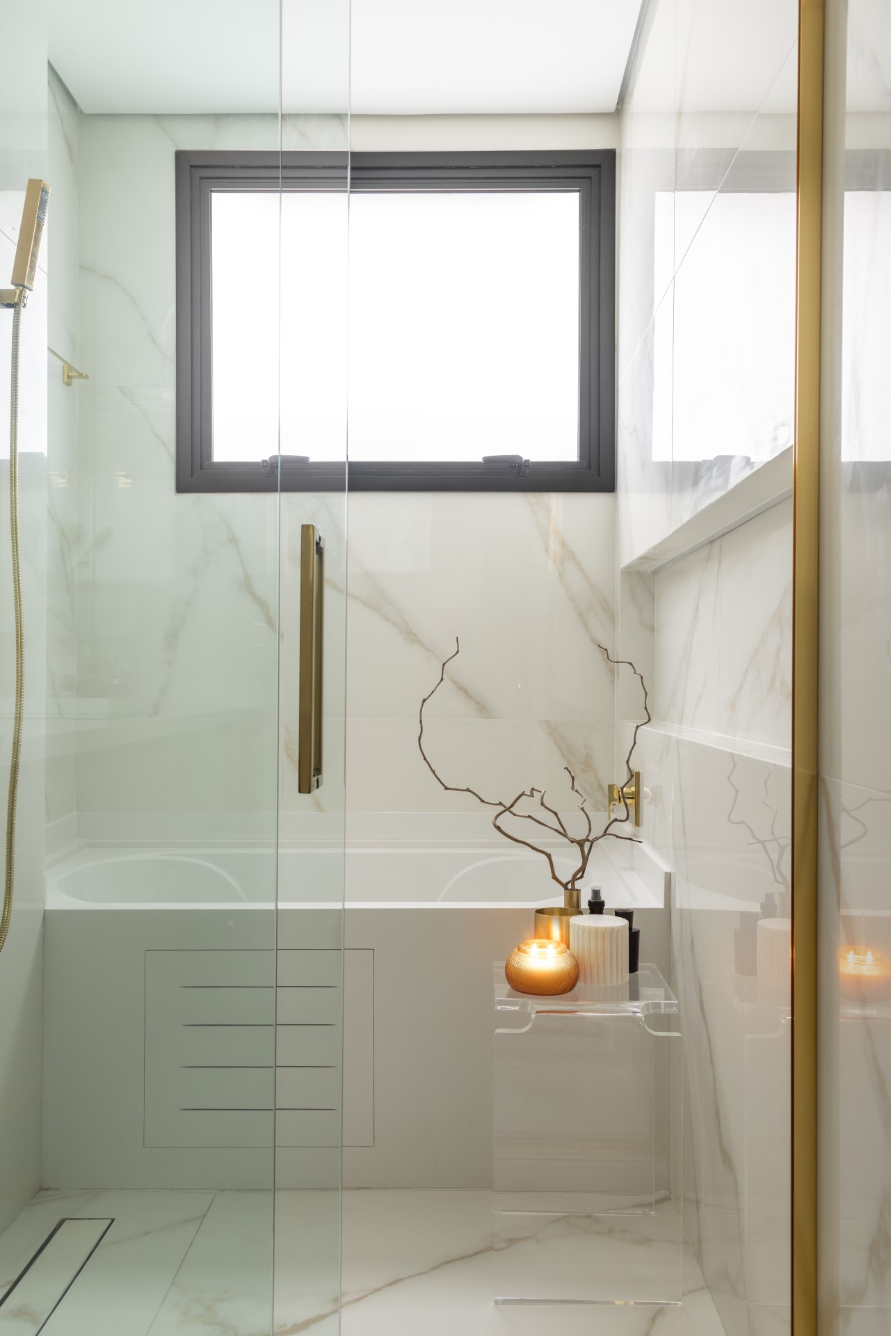 Projeto de Denise Polverini e Fernanda Villefort. Na foto, banheiro com revestimento de porcelanato marmorizado, banheira e metais dourados. Banco de acrílico ao lado da banheiro.