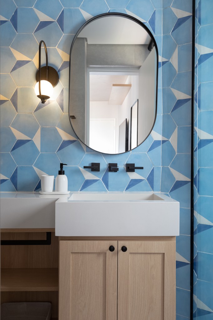 Banheiro pequeno com paredes de ladrilhos azuis, bancada branca, metais pretos e espelho oval.