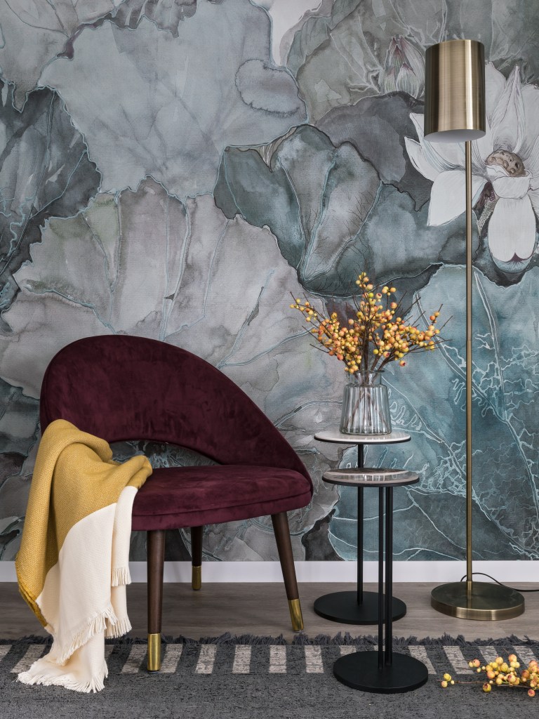 Quarto com parede com painel floral azulado, poltrona vinho e mesa lateral.