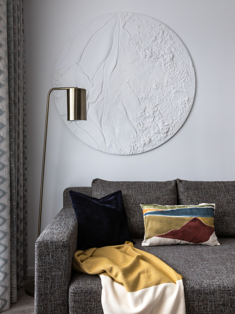 Sala de estar com sofá cinza escuro, luminária de piso dourada e obra de arte em relevo na parede.