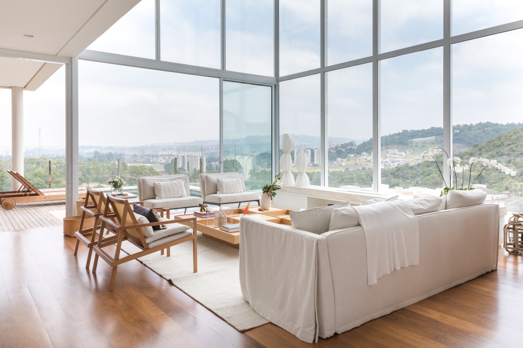 Sala de estar com paleta de cores claras, janelas grandes, pé-direito alto, piso de madeira e sofás brancos com poltronas.