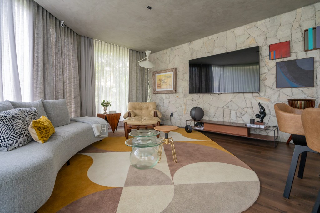 Projeto de Romário Rodrigues Arquitetos. Na foto, sala de estar com tapete geométrico, poltrona mole, janela curva com cortina e parede de pedras.