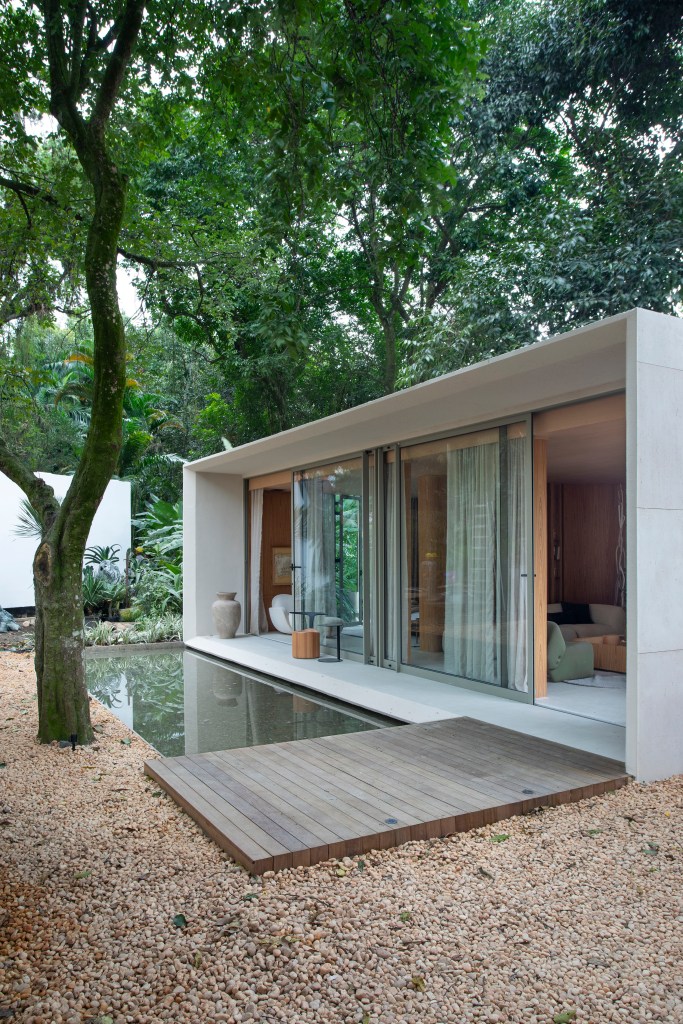 Casa de 90 m² é totalmente integrada à natureza com paredes de vidro. Projeto de Gabriela Eloy para a CASACOR Rio 2023. Na foto, fachada da casa com espelho d'água e jardim.