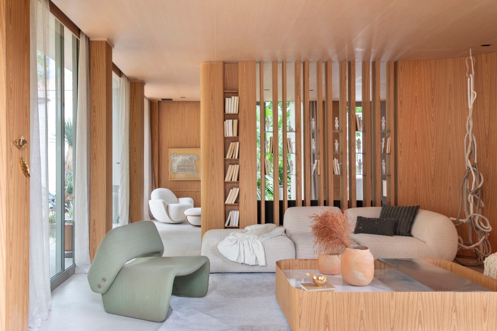 Casa de 90 m² é totalmente integrada à natureza com paredes de vidro. Projeto de Gabriela Eloy para a CASACOR Rio 2023. Na foto, sala da casa com brises de madeira e sofá curvo.