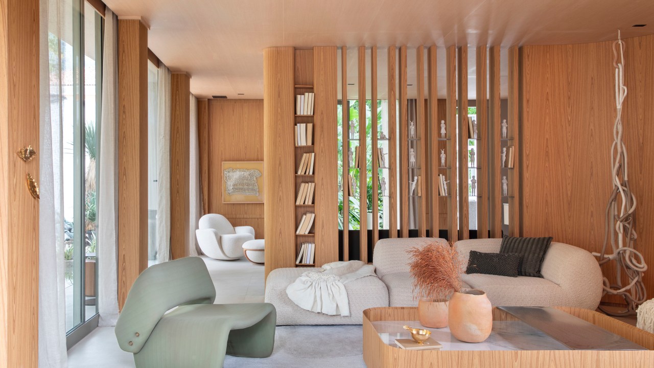 Casa de 90 m² é totalmente integrada à natureza com paredes de vidro. Projeto de Gabriela Eloy para a CASACOR Rio 2023. Na foto, sala da casa com brises de madeira e sofá curvo.