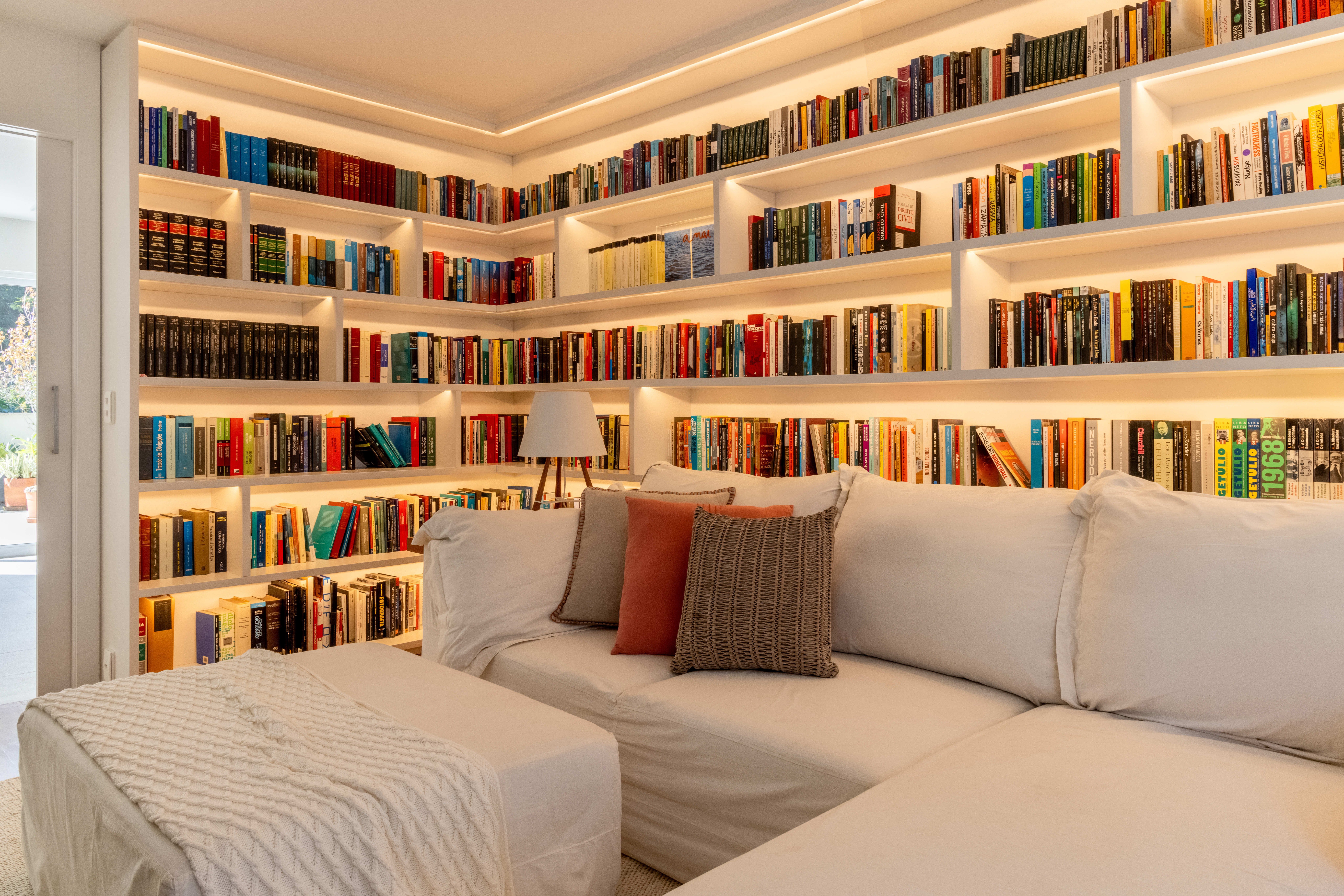 Projeto de Manuela Santos. Na foto, biblioteca com sofá branco L, estante retro iluminada com livros.