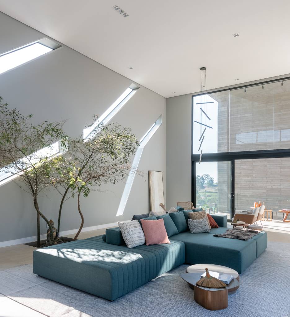 Sala de estar com pé-direito alto, sofá ilha azul e árvore.