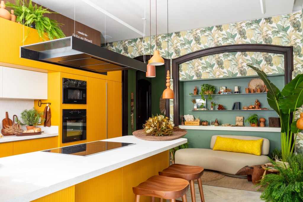 Cozinha com papel de parede de bananeira é repleta de artesanato brasileiro. Projeto de Amanda Miranda para a CASACOR Rio 2023. Na foto, cozinha com bancada de refeições, sofá e marcenaria amarela.