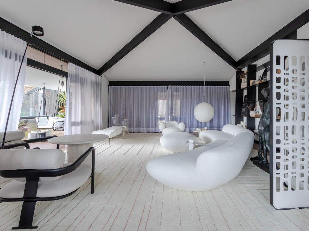 Living minimalista de 85 m² ganha décor em branco, preto, cinza e areia. Projeto de David Bastos para a CASACOR Bahia. Na foto, sala com sofá curvo branco, cortina, poltrona e marcenaria preta,