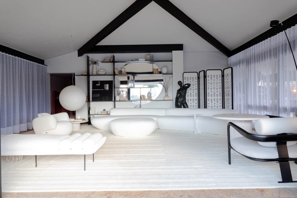 Living minimalista de 85 m² ganha décor em branco, preto, cinza e areia. Projeto de David Bastos para a CASACOR Bahia. Na foto, sala de estar com sofá curvo, estante preta, espelho e tapete.