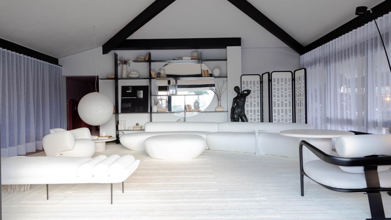 Living minimalista de 85 m² ganha décor em branco, preto, cinza e areia. Projeto de David Bastos para a CASACOR Bahia. Na foto, sala de estar com sofá curvo, estante preta, espelho e tapete.
