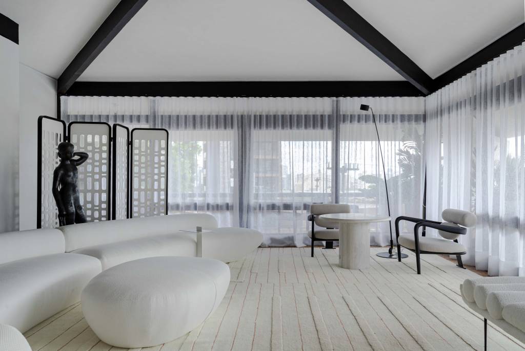 Living minimalista de 85 m² ganha décor em branco, preto, cinza e areia. Projeto de David Bastos para a CASACOR Bahia. Na foto, sala de estar com sofá curvo, estante preta, espelho e tapete.