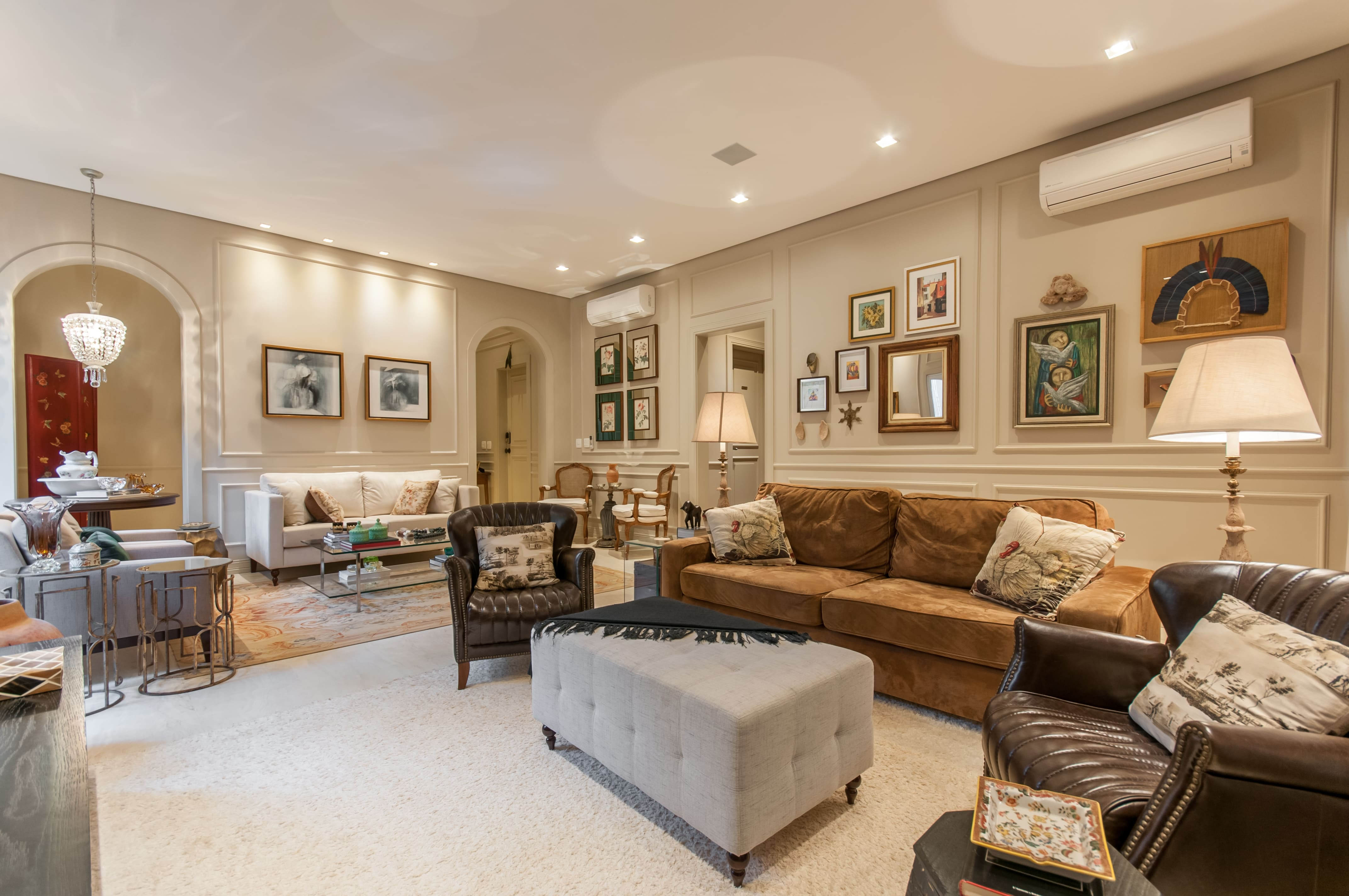 Sala de estar com decoração clássica, boiseries nas paredes, sofá marrom, poltronas de couro e pufe como mesa de centro.