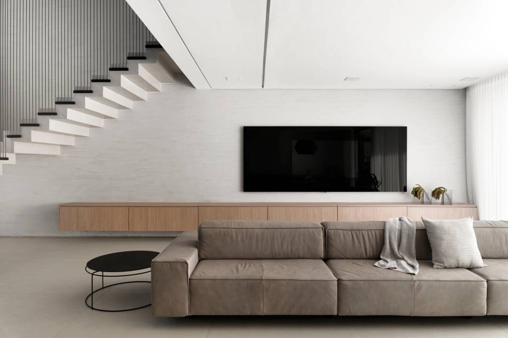 Sala de tv minimalista com parede cinza clara, rack de madeira e sofá baixo; escada branca com guarda corpo preto.