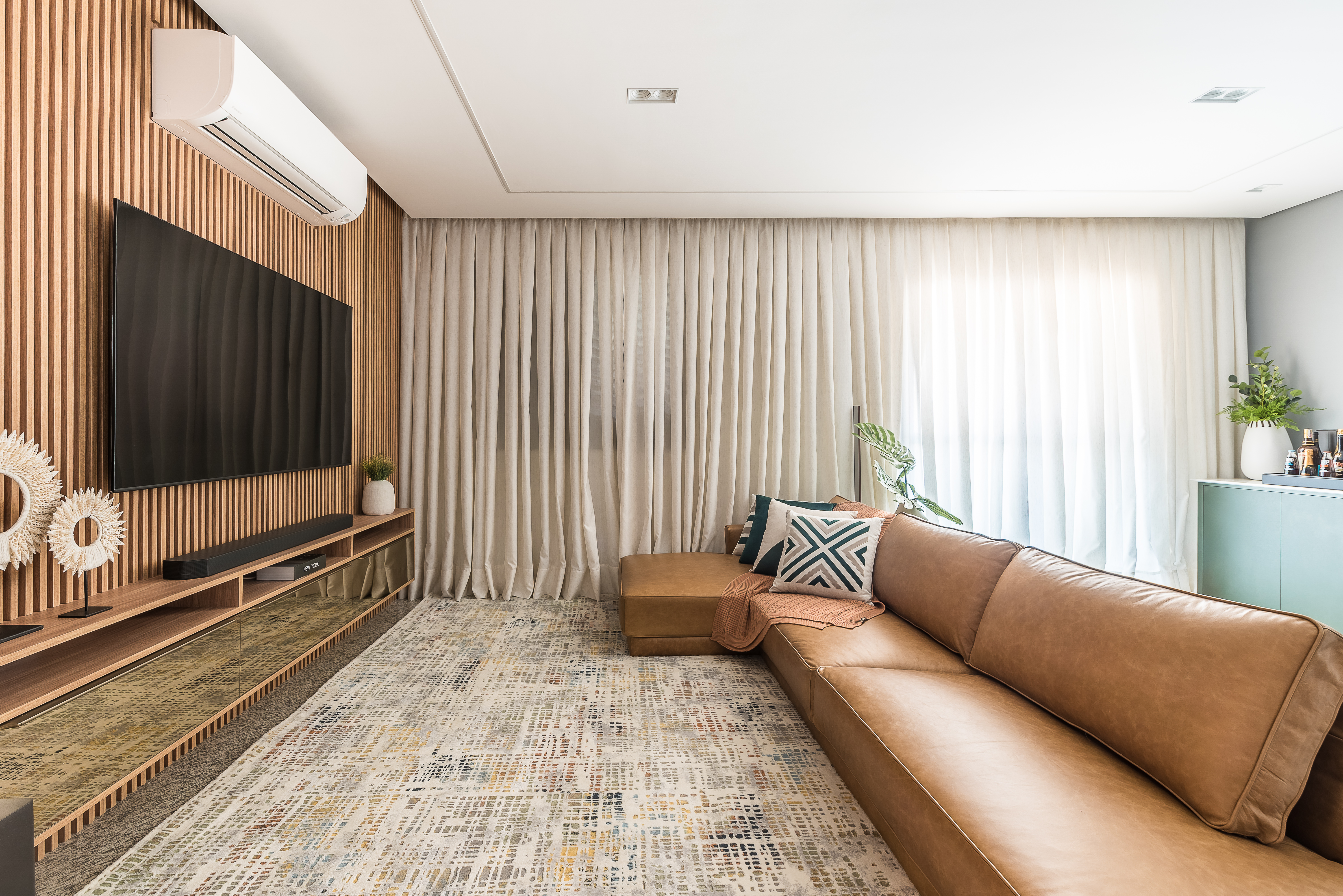 Projeto de Clareira Arquitetura. Na foto, sala de estar com painel ripado, sofá de couro e cortina.