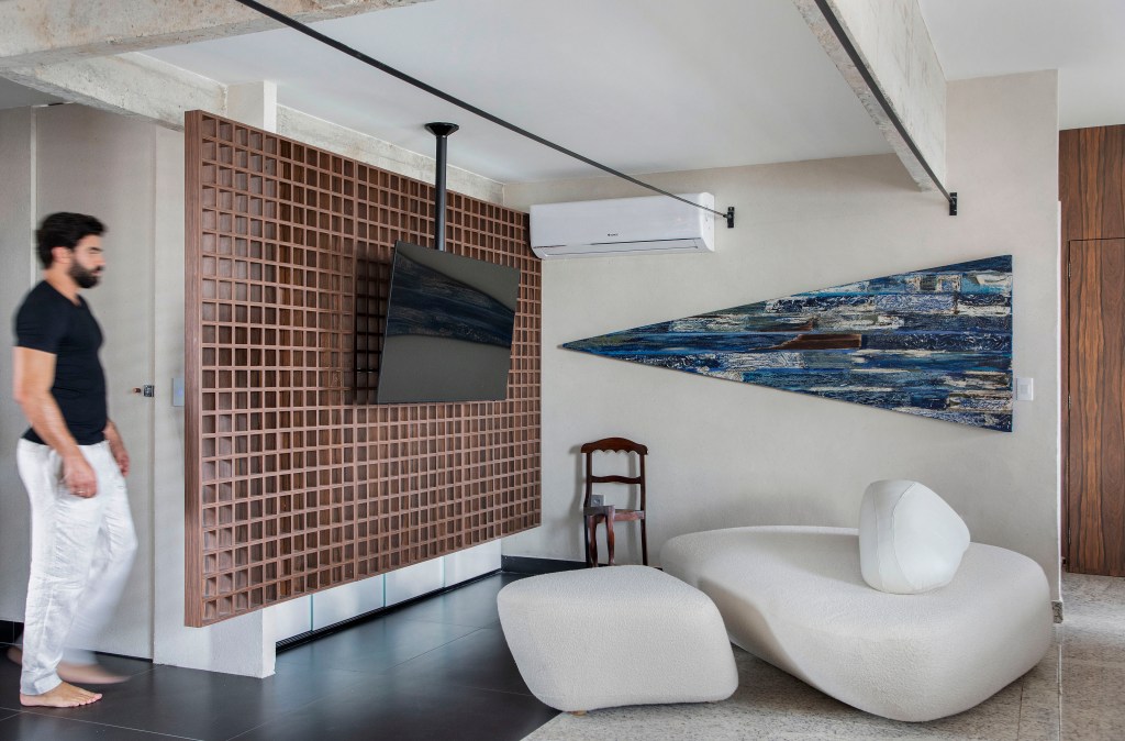 Projeto de Roby Macedo. Na foto, sala de estar com concreto aparente e sofá ilha em formato orgânico branco. Painel vazado em forma de muxarabi para tv.