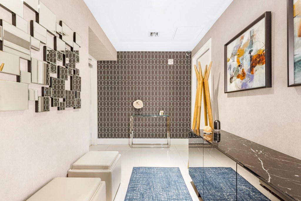 Hall de entrada com aparador espelhado, pufes brancos, tapete azul e parede com papel de parede geométrico preto ao fundo.