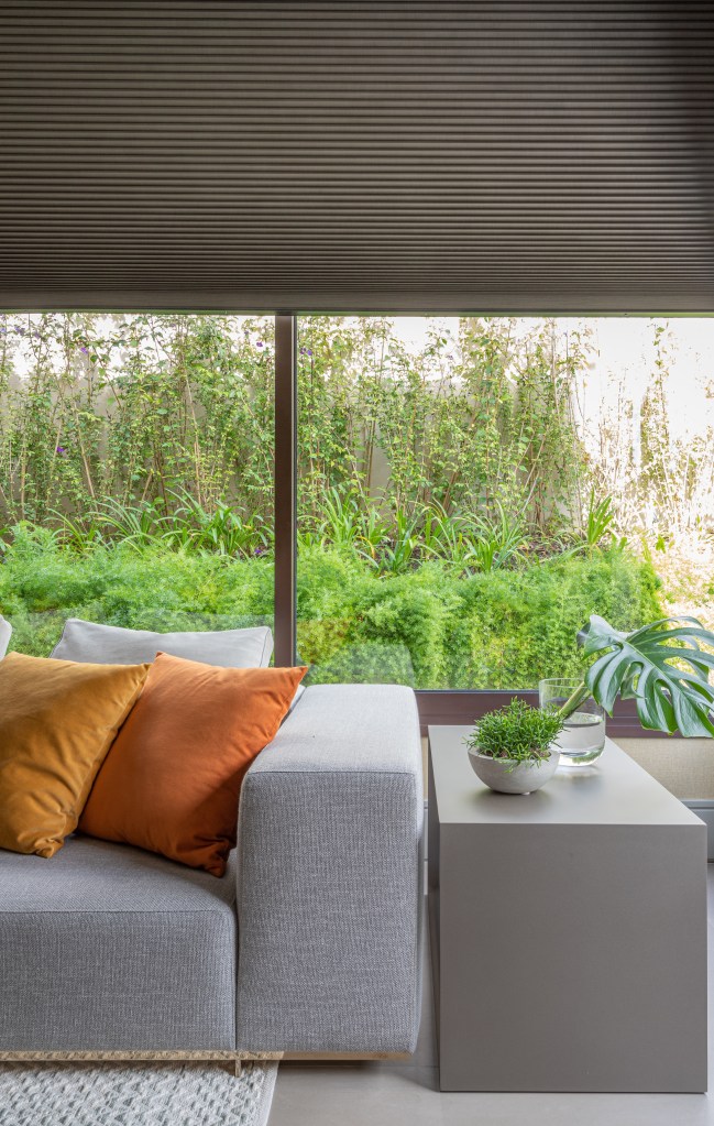 Projeto de Patricia Penna. Sala de estar com sofá cinza, mesa lateral, janela para jardim com persiana.
