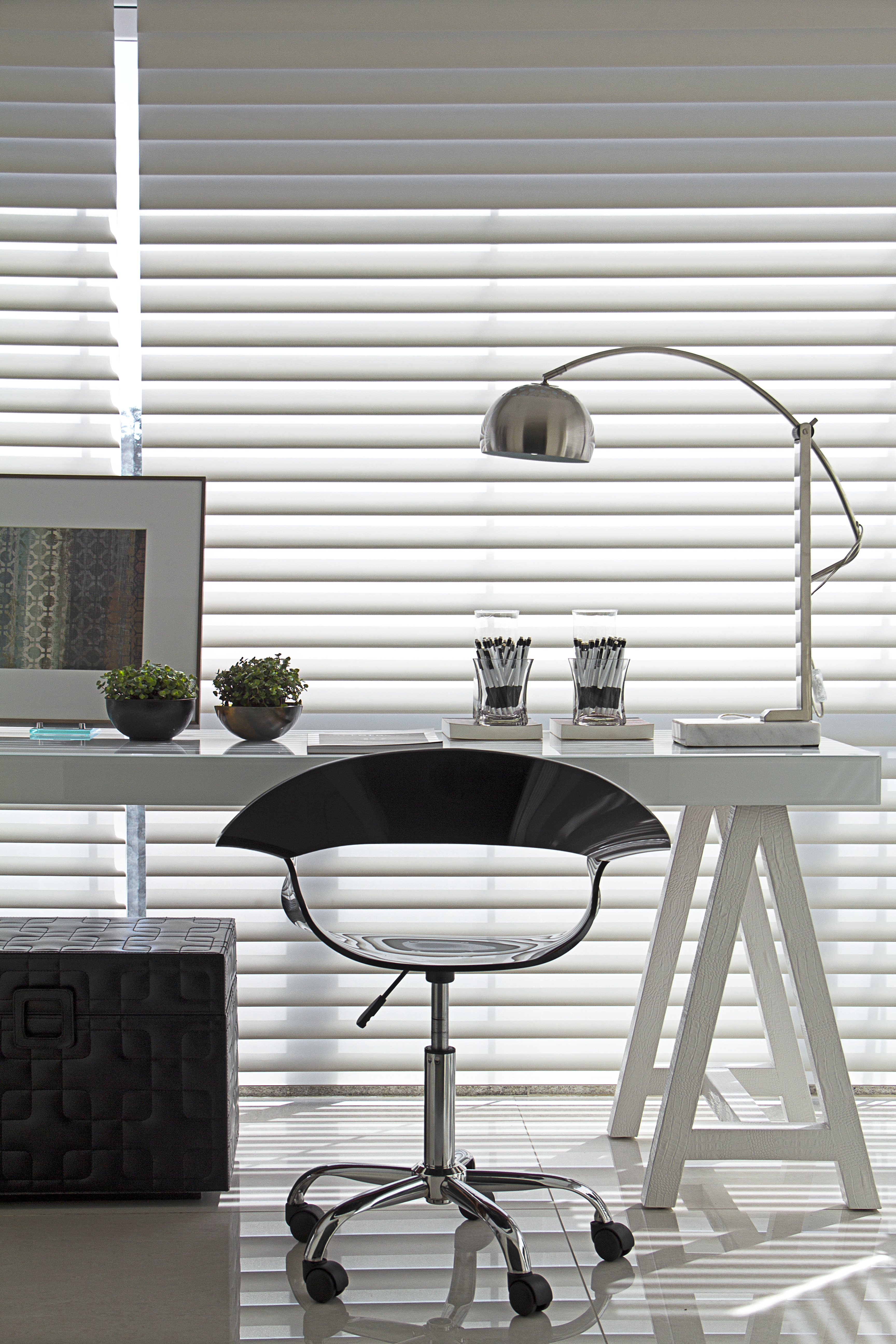 Projeto de Patricia Penna. Home office com mesa branca de cavaletes, cadeira preta e persiana branca.
