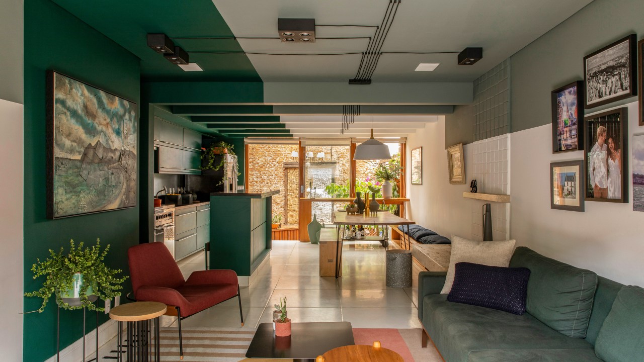 Projeto de Ricardo Abreu. Na foto, sala integrada com cozinha e sala de jantar. Teto verde e ilha da cozinha verde, tapete listrado e poltrona vermelha.