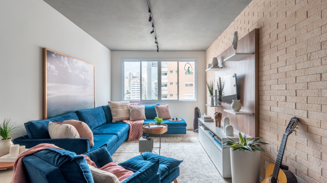 Projeto de Isabella Nalon. Na foto, sala com sofá azul em L, poltrona azul, iluminação com trilho de spot e parede de tijolinhos.