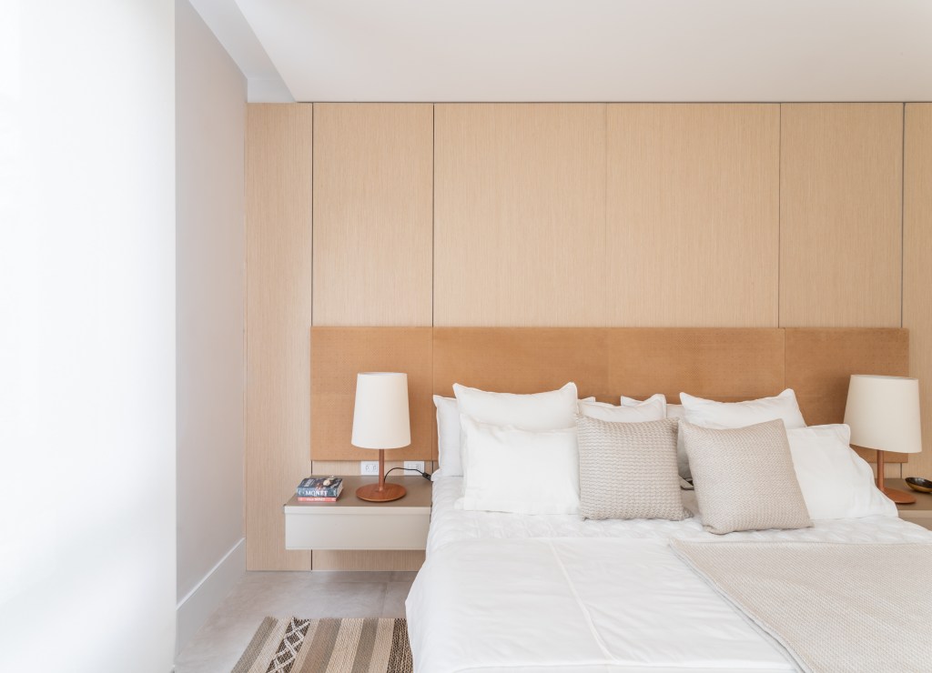 Quarto clean com cama de casal, cabeceira bege, tapete e parede revestida de madeira.