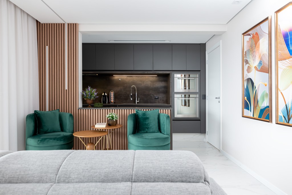 Projeto de Palladino Arquitetura. Na foto, cozinha integrada com marcenaria cinza, piso de porcelanato e sala com poltronas verdes.