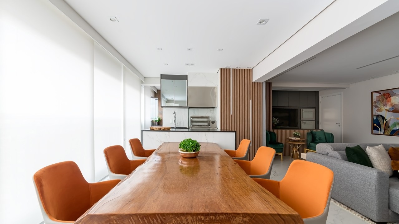 Projeto de Palladino Arquitetura. Na foto, varanda integrada com mesa grande de madeira e cadeiras com estofado laranja.