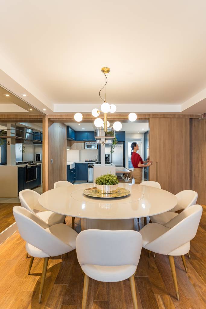 Sala de jantar com mesa redonda e cadeiras brancas. Cozinha com porta de correr ao fundo.