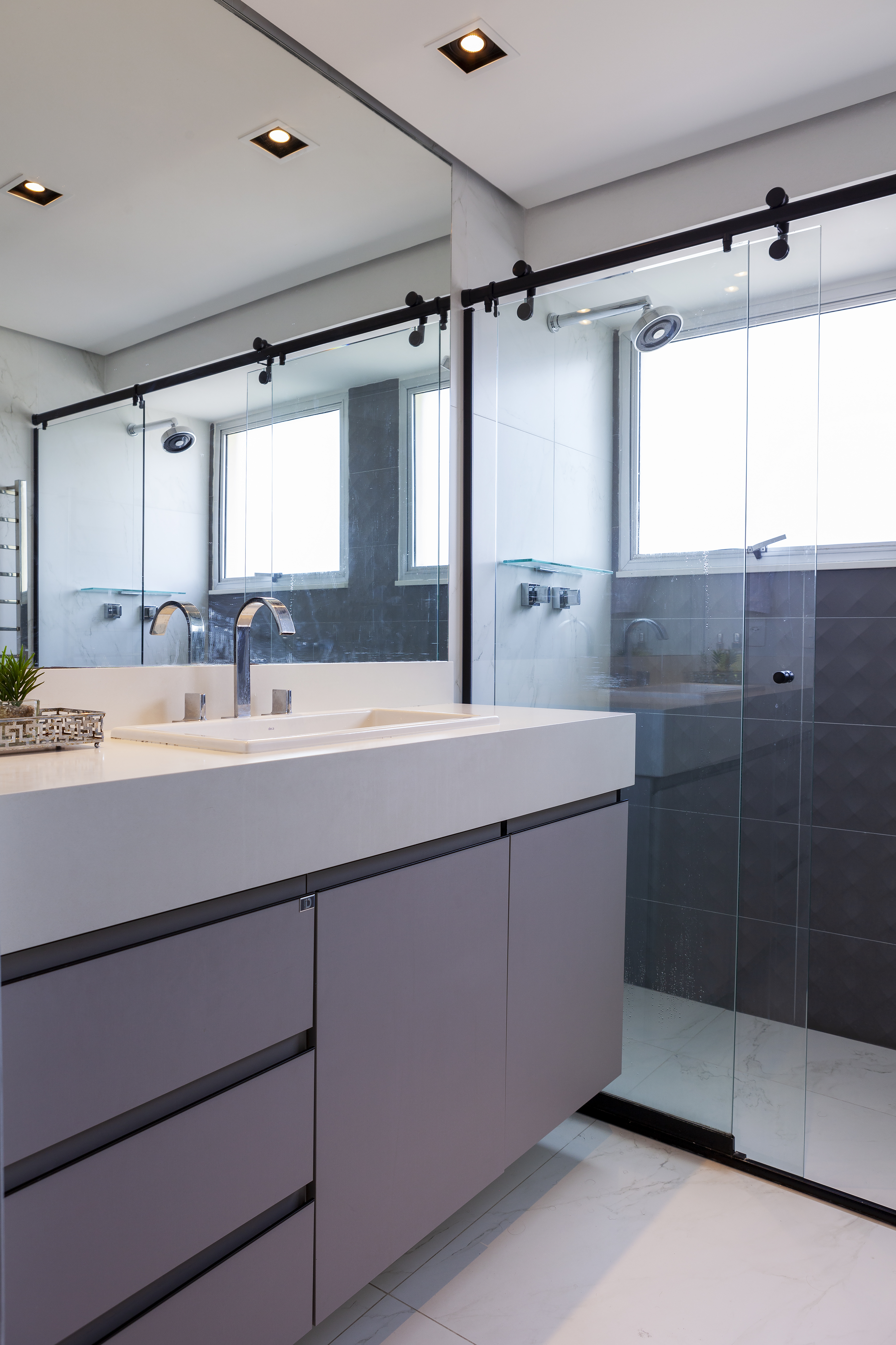 Banheiro com bancada branca, marcenaria cinza e box de vidro.