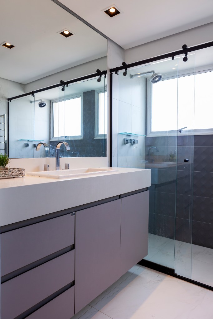 Banheiro com bancada branca, marcenaria cinza e box de vidro.