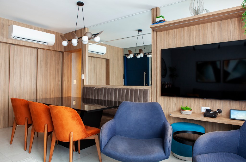 Sala de estar com poltrona azul, mesa de jantar com banco e cadeiras laranja, espelho.