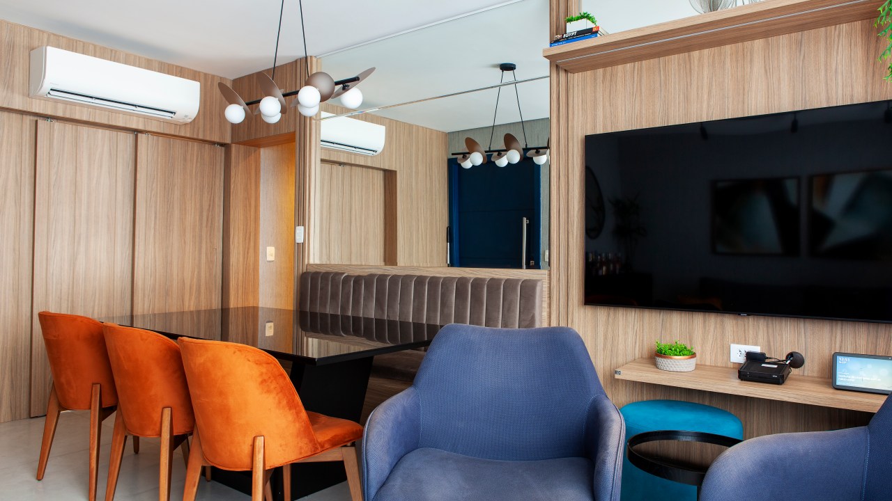 Sala de estar com poltrona azul, mesa de jantar com banco e cadeiras laranja, espelho.