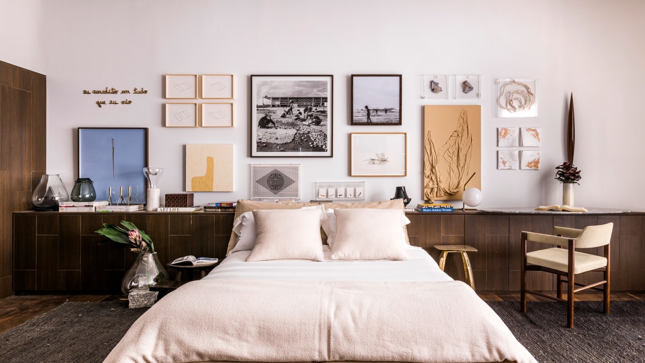 Quarto com cama de casal e bancada de madeira escura com cadeira; quadros na parede.