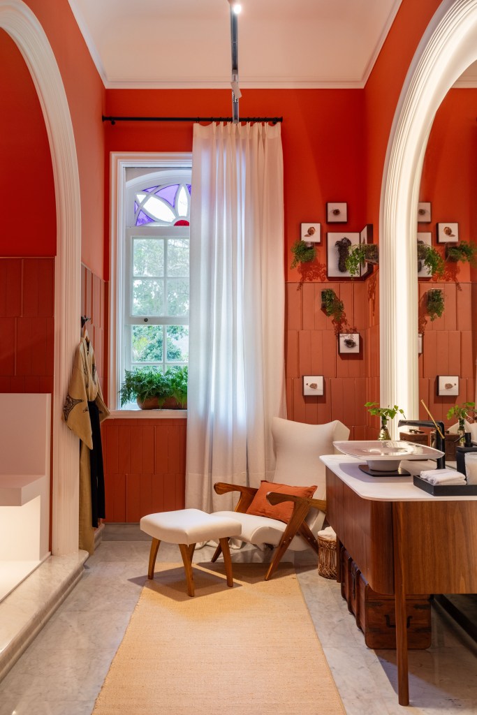 Suíte ganha conforto quarto de hotel com paredes de madeira. Projeto de Paula Muller para a CASACOR Rio 2023. Na foto, banheiro com parede laranja e espelho.