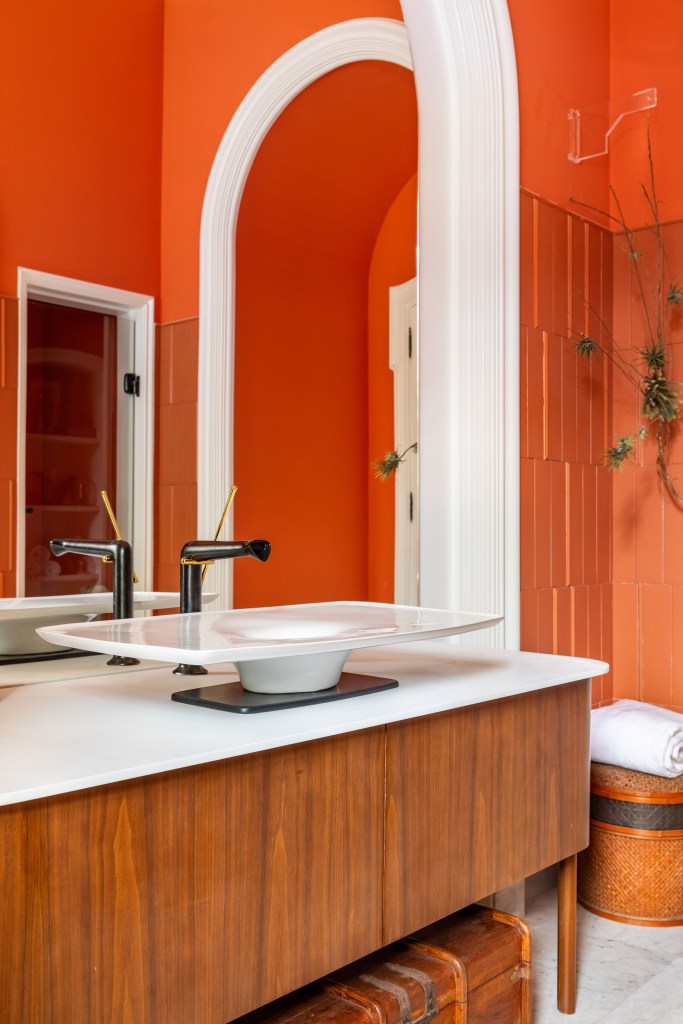 Suíte ganha conforto quarto de hotel com paredes de madeira. Projeto de Paula Muller para a CASACOR Rio 2023. Na foto, banheiro com parede laranja e espelho.